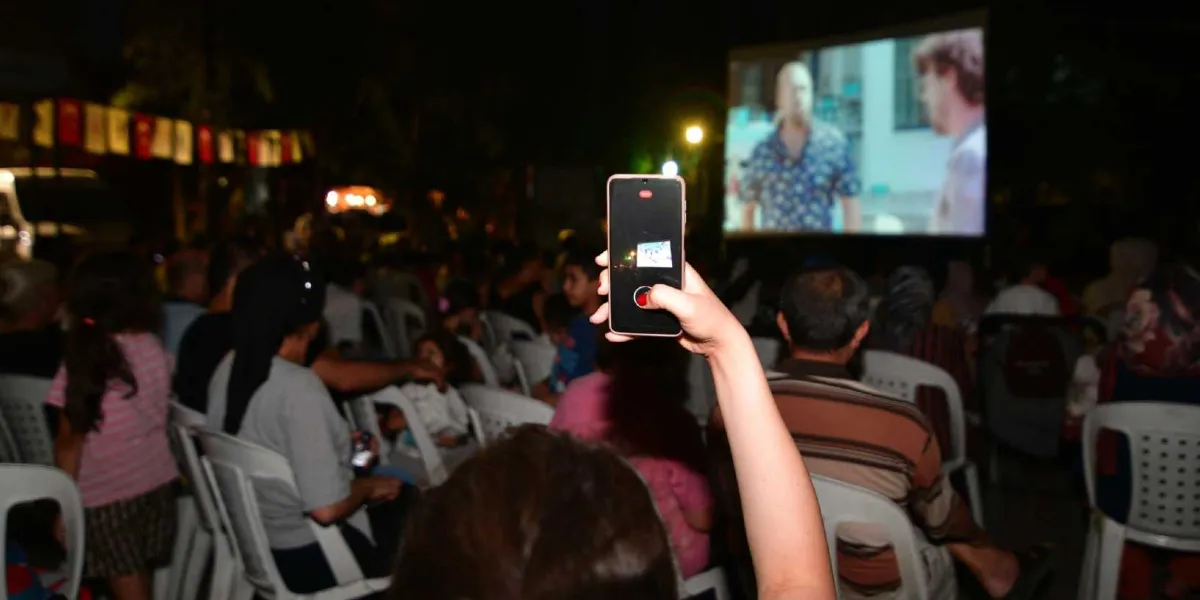 Gece vakti açık havada kurulan bir sinema gösteriminde, izleyiciler beyaz plastik sandalyelerde oturarak perdeye yansıtılan filmi izliyor. Ön planda bir izleyicinin elinde tuttuğu akıllı telefon, ekranı üzerinden sinema perdesini kaydediyor. Perdede iki erkek karakterin yer aldığı bir film sahnesi görülüyor. Arka planda ise ağaçlar ve etkinlik alanını çevreleyen süs bayrakları seçiliyor.