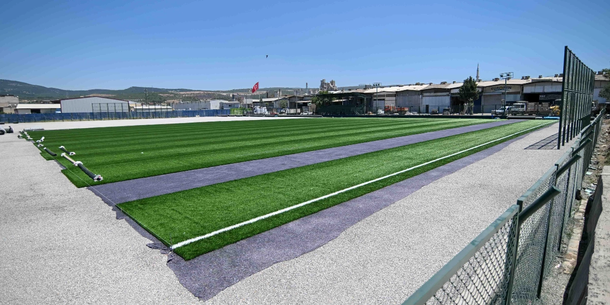 Yapım aşamasında olan, zemini yeni serilmiş yeşil suni çimle kaplı bir futbol sahası. Saha kenarında henüz döşenmemiş çim ruloları ve siyah zemin kaplama malzemeleri görülüyor. Arka planda sanayi bölgesi yapıları, bir cami minaresi ve tepelik bir arazi yer alıyor.