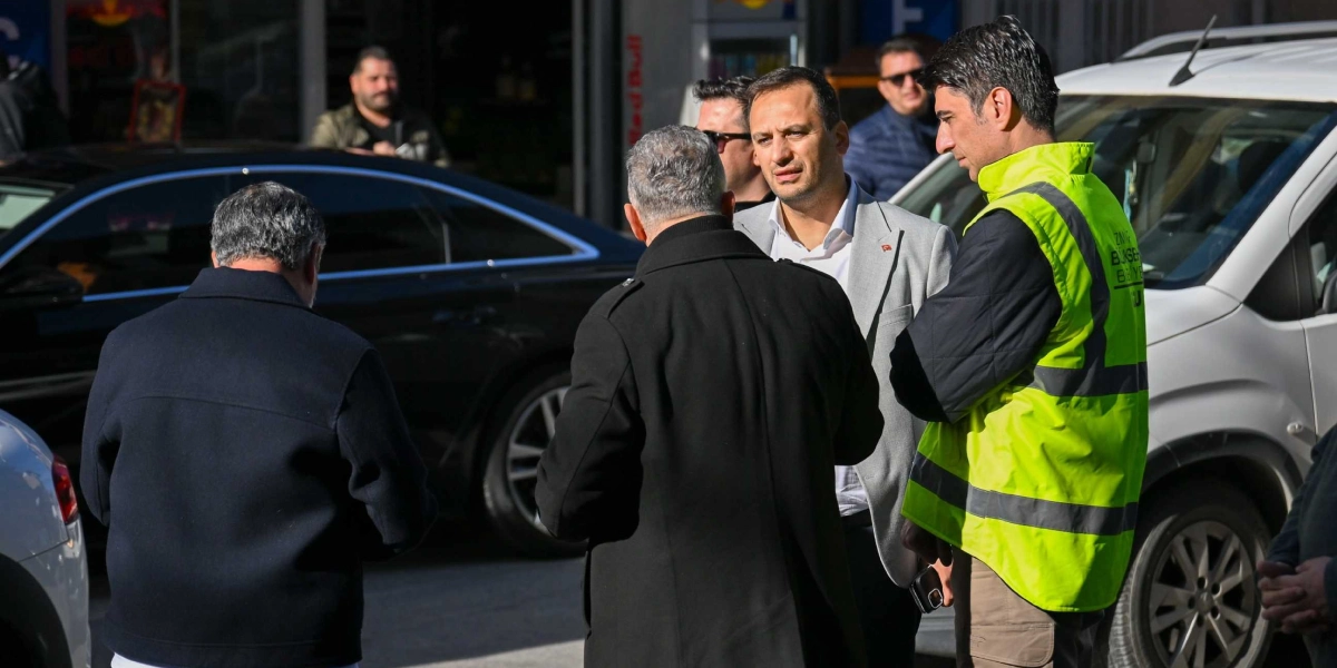 Bornova Belediye Başkanı Mustafa İduğ ve beraberindeki heyet, bir çalışma sahasında incelemelerde bulunuyor. Başkan İduğ, üzerinde gri bir ceket ve beyaz gömlek ile bir grup yetkiliyle sohbet ederken, yanında üzerinde 
