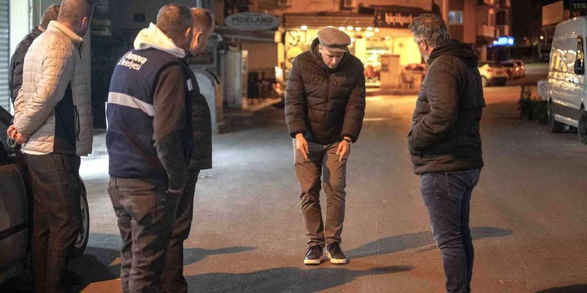 Gece vakti bir sokakta toplanmış, üzerinde Bornova Belediyesi yazılı yelek giyen bir görevlinin de bulunduğu beş kişilik bir grup, yerde yeni yapılmış bir asfalt çalışmasını incelemektedir. Grubun ortasında yer alan kasketli bir kişi, asfalt üzerindeki bir noktayı işaret ederek diğerlerine göstermektedir.
