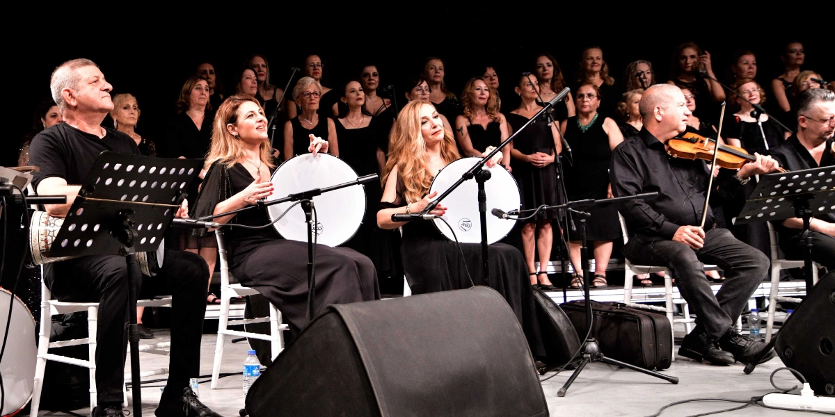 Siyah kıyafetler giymiş bir müzik grubu ve arkalarında duran koro, sahnede konser veriyor. Ön planda oturan müzisyenlerden soldaki elinde tef tutuyor, yanındaki kadın da tef çalıyor, sağ taraftaki müzisyen ise keman çalıyor. Müzisyenlerin önünde nota sehpaları ve mikrofonlar bulunuyor. Arka planda ise ayakta duran kadınlardan oluşan kalabalık bir koro yer alıyor.