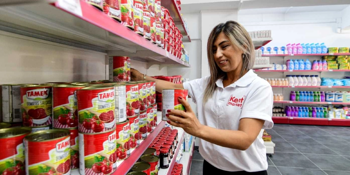 Üzerinde Kent Market yazılı beyaz bir tişört giyen kadın çalışan, market rafındaki Tamek marka domates salçası konservelerini düzenliyor. Raflarda çok sayıda konserve kutusu ve arka planda temizlik ürünlerinin bulunduğu diğer market reyonları yer alıyor.