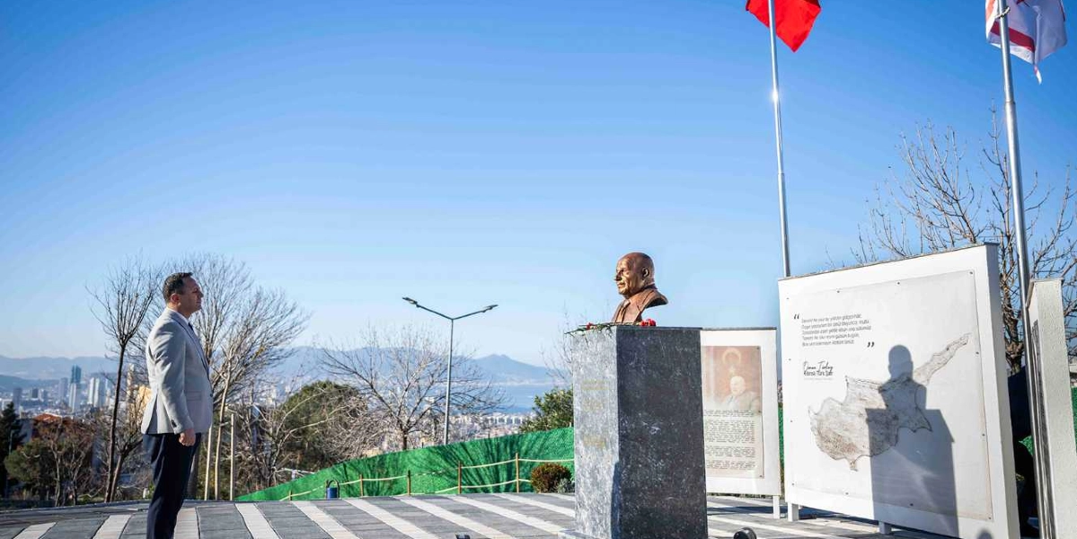 Açık havada, Bornova manzarasına karşı konumlanmış Rauf Denktaş büstünün önünde saygı duruşunda bulunan bir erkek. Büstün yanında, üzerinde Kıbrıs haritası ve Denktaş'ın bir sözünün yer aldığı bilgilendirme panoları ile Türkiye ve Kuzey Kıbrıs Türk Cumhuriyeti bayrakları bulunuyor.