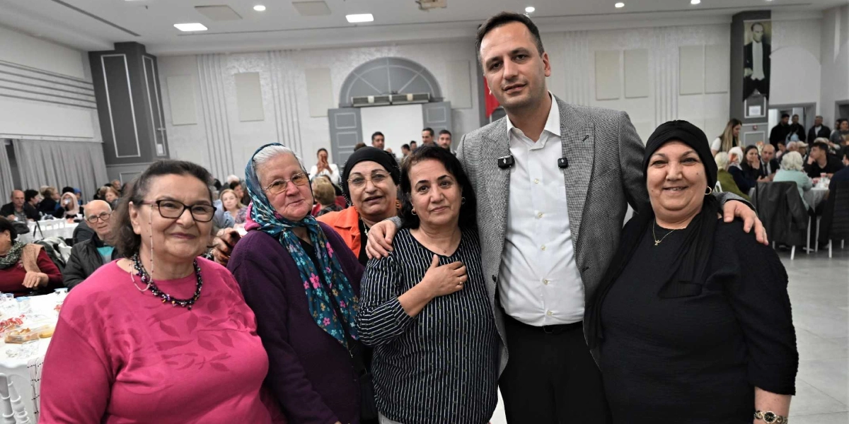 Geniş ve aydınlık bir salonda düzenlenen dayanışma yemeğinde, gri ceketli bir erkek, yan yana duran dört kadınla birlikte gülümseyerek poz veriyor. Kadınlar günlük kıyafetler ve başörtüleri içinde, samimi bir ortamda bir araya gelmiş durumda. Arka planda, masalarda oturan diğer davetliler ve salonun genel düzeni görülüyor.