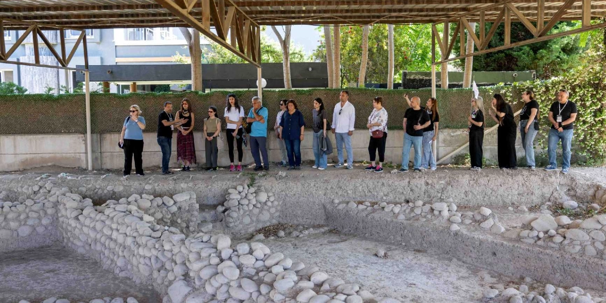 Üstü metal bir çatı ile kapatılmış açık hava arkeolojik kazı alanında, bir grup insan rehberi dinleyerek tarihi kalıntıları inceliyor. Taş duvar temellerinin bulunduğu kazı alanının hemen arkasında, yeşil çitlerle çevrili bir bahçe ve ağaçlar yer alıyor.