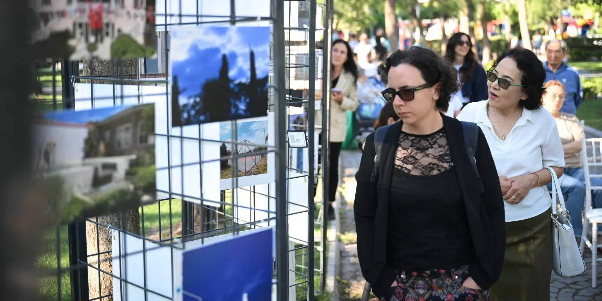 Açık havada kurulan metal bir sergileme panosunda asılı fotoğrafları inceleyen insanlar. Ön planda siyah hırkalı ve güneş gözlüklü bir kadın ile yanında beyaz bluzlu, gözlüklü başka bir kadın fotoğraflara bakarak yürüyor. Arka planda ise sergiyi gezen diğer ziyaretçiler ve ağaçlıklı bir park alanı görülüyor.