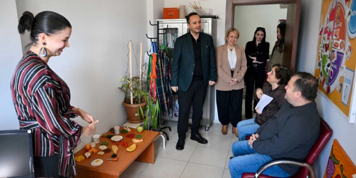 Ofis ortamında, masanın üzerinde çeşitli besin maketlerinin bulunduğu bir sunum sırasında, ayakta duran bir kadın katılımcılara bilgi veriyor. Masanın karşısında oturan bir erkek ve bir kadın ile ayakta duran diğer üç kişi, konuşmacıyı dinleyerek sohbet ediyor. Duvarın bir kısmında besin gruplarını gösteren eğitici bir pano asılı bulunuyor.