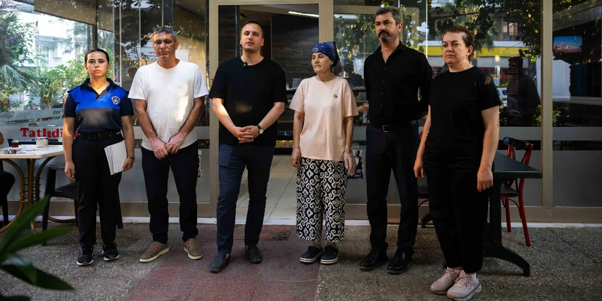 Bir fırın veya işletmenin önünde yan yana duran altı kişilik bir grup; soldan sağa doğru zabıta üniformalı bir kadın, beyaz tişörtlü bir erkek, siyah tişörtlü bir erkek, başında mavi bir bandana olan açık renkli tişörtlü bir kadın, siyah gömlekli bir erkek ve siyah tişörtlü bir kadın yer alıyor. Grup, denetim çalışmaları kapsamında işletme önünde durarak objektiflere bakıyor.
