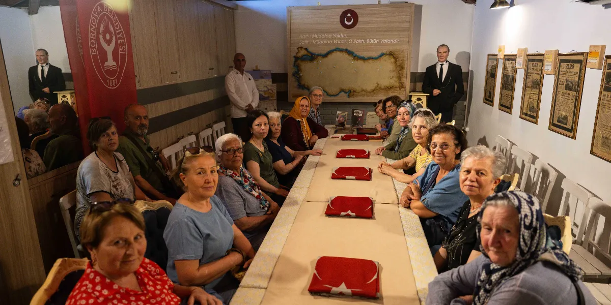 Bornova Belediyesi'ne ait bir müze veya etkinlik alanında, uzun bir masa etrafında toplanmış çoğunluğu kadınlardan oluşan bir grup ziyaretçi. Masanın üzerinde Türk bayrağı motifli örtüler ve Nutuk kitabı bulunuyor. Arka planda duvarda Türkiye haritası üzerinde 