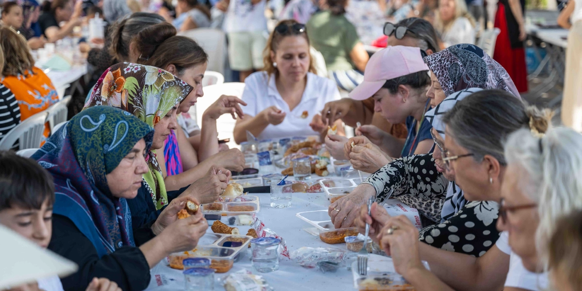 Açık havada uzun bir masanın etrafında toplanmış, plastik tabaklarda menemen ve simit yiyen kadınlar ve çocuklar. Katılımcıların çoğu başörtülü olup, samimi bir ortamda hep birlikte yemek yiyorlar.