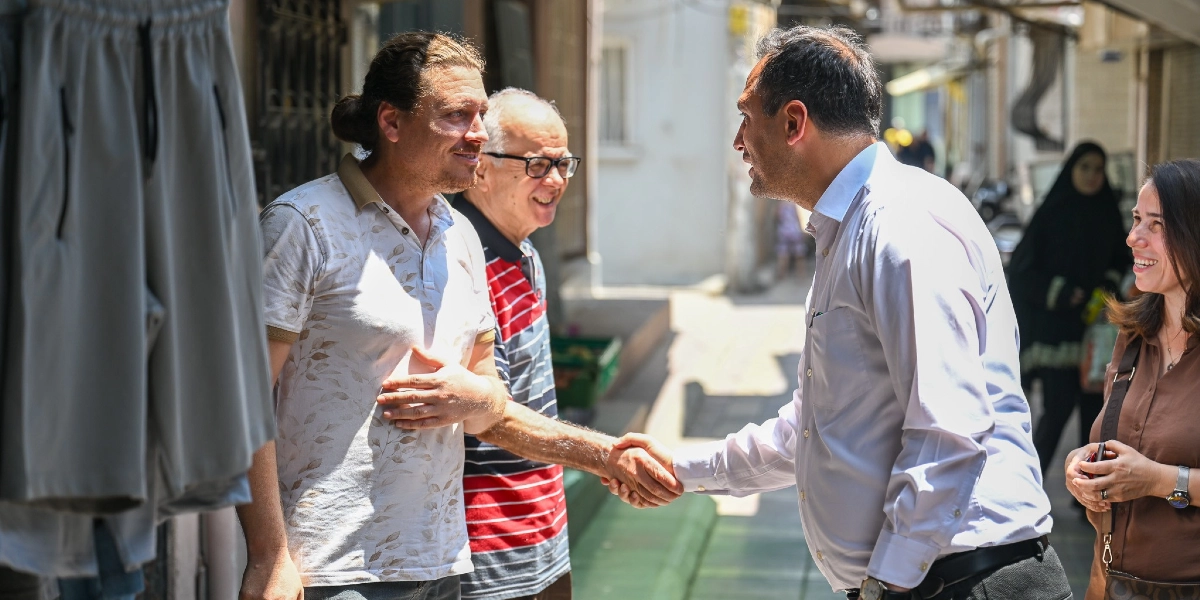 Güneşli bir sokakta, açık renkli gömlek giyen bir erkek, esnaf olduğu anlaşılan saçları arkadan bağlı, desenli tişörtlü bir adamla el sıkışıyor. Yanlarında gözlüklü, çizgili tişörtlü yaşlı bir adam ve gülümseyerek onları izleyen kahverengi bluzlu bir kadın bulunuyor. Ön planda asılı duran gri şortlar kısmen görünüyor.