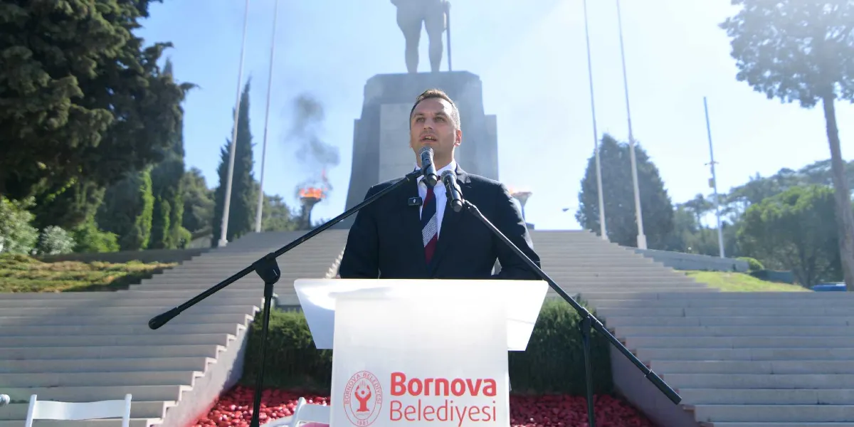 Bornova Belediye Başkanı, üzerinde Bornova Belediyesi logosu bulunan bir kürsüde, arkasında Atatürk anıtı ve yanan meşalelerin olduğu bir tören alanında konuşma yapıyor.