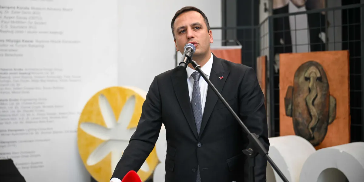 Siyah takım elbiseli, beyaz gömlekli ve gri kravatlı bir erkek, önündeki mikrofona konuşma yapıyor. Yaka kısmında küçük bir Türk bayrağı rozeti bulunan kişinin arkasında, duvarda isimlerin yazılı olduğu bir pano ve sağ tarafta turuncu renkli, üzerinde soyut bir figür bulunan bir tablo yer alıyor. Arka planda ayrıca sarı renkli, çiçek formunda büyük bir dekoratif obje ve beyaz silindirik yapılar bulunuyor.