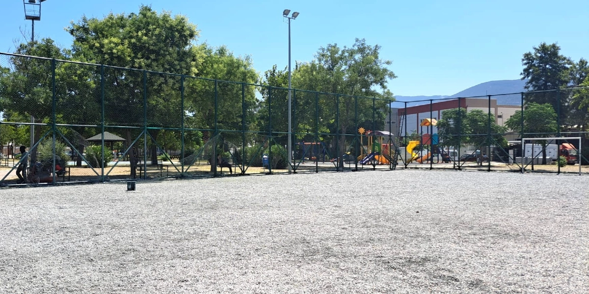 Açık havada, zemini gri çakıl taşlarıyla kaplı, etrafı yüksek tel örgülerle çevrili bir futbol sahası. Sahasının arka planında ağaçlar, bir çocuk parkı oyun grubu ve uzakta dağ manzarası görülüyor.