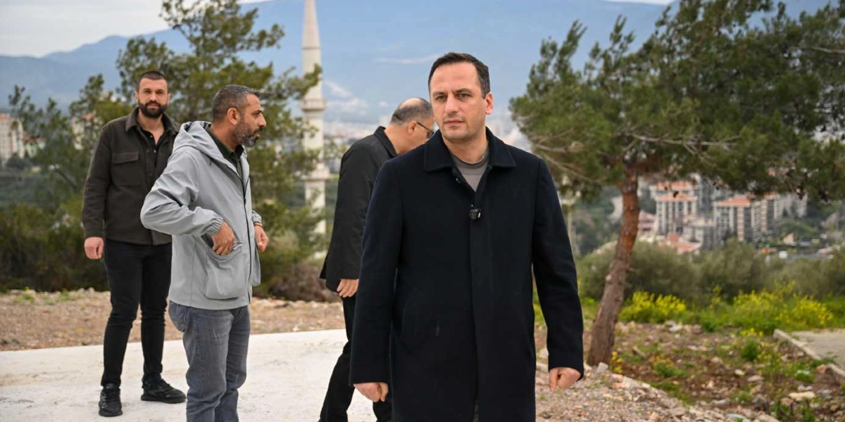Bornova Belediye Başkanı Ömer Eşki, beraberindeki heyetle birlikte İZYUVA bölgesindeki arazide incelemelerde bulunuyor. Arka planda ağaçlık bir alan, bir cami minaresi ve uzaktan görünen yerleşim yerleri yer alıyor.