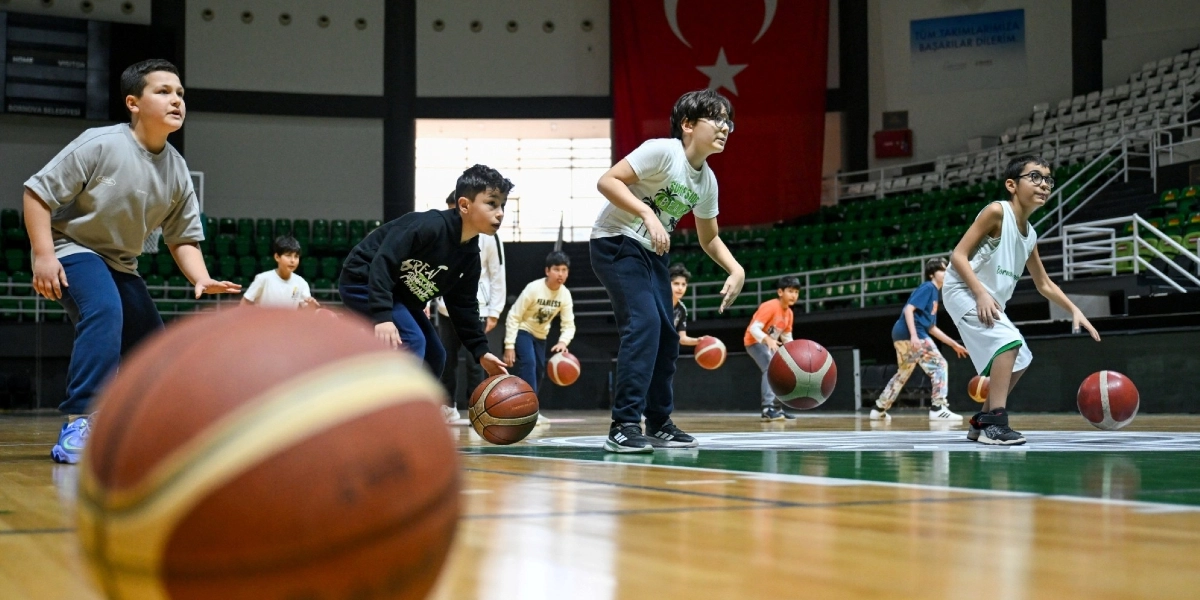 Kapalı bir spor salonunda basketbol antrenmanı yapan bir grup çocuk, ellerinde basketbol toplarıyla sahada sıralanmış şekilde duruyor. Ön planda odak dışı büyük bir basketbol topu yer alırken, arka planda duvarda asılı büyük bir Türk bayrağı ve boş tribünler görülüyor. Çocuklar spor kıyafetleri içinde, dikkatli bir şekilde top sürme çalışması yapıyor.