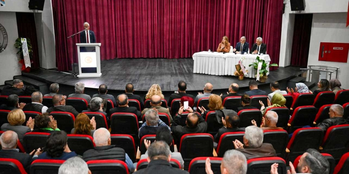 Konferans salonunda kürsüde konuşma yapan bir kişi ve onu dinleyen kalabalık bir grup muhtar yer alıyor. Sahnenin arka planında bordo renkli perdeler bulunurken, sağ taraftaki masada üç kişi oturuyor. İzleyiciler, kırmızı koltuklarda oturarak konuşmacıyı dinliyor ve bazıları alkışlıyor.