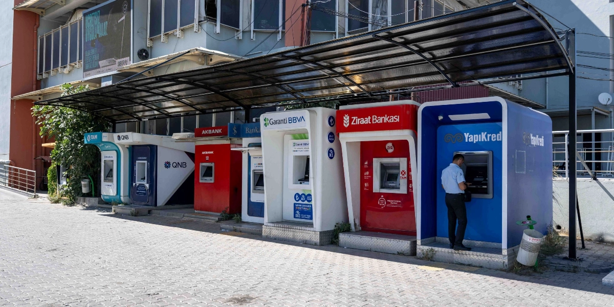 Bornova'da güneşten ve yağmurdan korunmak için üzerleri ortak bir çatı ile kapatılmış yan yana dizili altı farklı bankaya ait ATM kabinleri. Soldan sağa doğru QNB, Akbank, Garanti BBVA, Ziraat Bankası ve Yapı Kredi ATM'leri sıralanıyor. En sağdaki Yapı Kredi ATM'sini kullanan bir kişi bulunuyor.