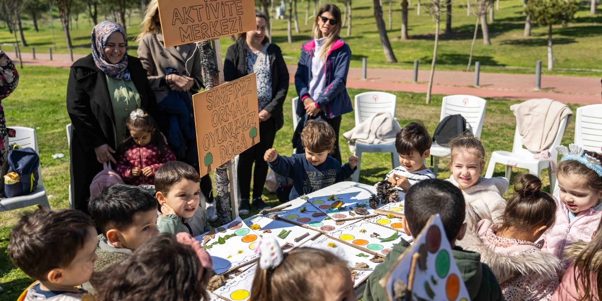 Açık havada, yeşil bir park alanında kurulan masanın etrafında toplanmış çocuklar ve onlara eşlik eden yetişkinler. Çocuklar, üzerinde renkli daireler ve doğadan toplanmış yaprak, dal, kozalak gibi materyallerin bulunduğu kağıtlarla 