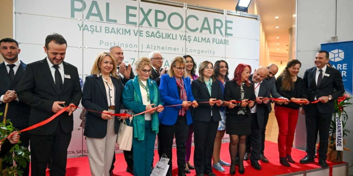 PAL EXPOCARE Yaşlı Bakım ve Sağlıklı Yaş Alma Fuarı'nın açılış kurdelesini kesen, aralarında Bornova protokolünden isimlerin de bulunduğu bir grup kadın ve erkek katılımcı, fuar standının önünde yan yana dizilmiş şekilde duruyor. Arka planda fuarın adının ve odaklandığı Parkinson, Alzheimer ve uzun yaşam gibi konuların yazılı olduğu büyük bir panel yer alıyor.