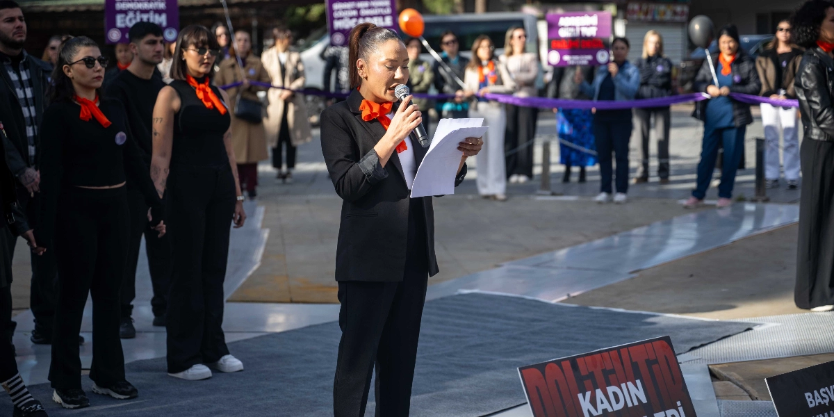 Açık havada düzenlenen bir eylemde, siyah ceketli bir kadın elinde mikrofon ve bir metinle konuşma yapıyor. Konuşmacının arkasında, ellerinde mor kurdeleler tutan ve boyunlarında turuncu fularlar takan bir grup kadın ve erkek destekçi yer alıyor. Arka planda üzerinde 