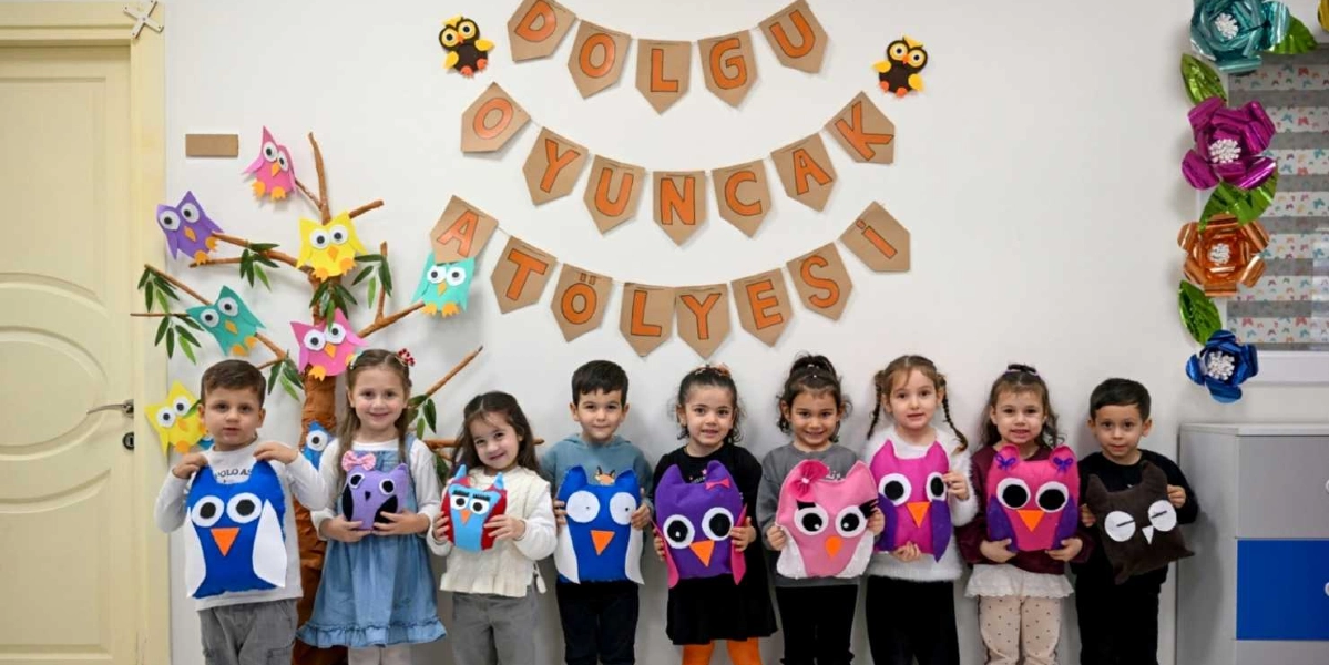 Beyaz bir duvarın önünde, ellerinde kendi yaptıkları renkli keçe baykuş oyuncaklarını tutan dokuz çocuk yan yana duruyor. Duvarın üzerinde karton harflerle 