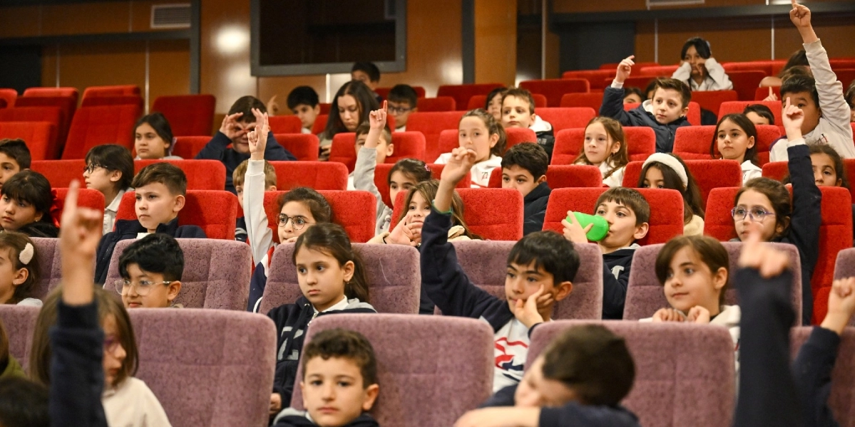 Konferans salonunda kırmızı koltuklarda oturan çok sayıda ilköğretim çağındaki öğrenci, bir etkinlik sırasında söz almak için ellerini havaya kaldırıyor. Bazı öğrenciler dikkatle dinlerken, bazıları ise el kaldırarak katılımcı bir tavır sergiliyor.