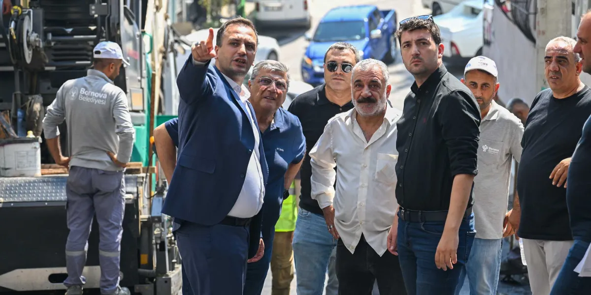 Bornova Belediye Başkanı Ömer Eşki, bir grup yetkiliyle birlikte asfalt serme çalışması yapılan bir sokakta incelemelerde bulunuyor. Lacivert takım elbiseli Başkan Eşki, eliyle çalışma alanını işaret ederek yanındakilere bilgi veriyor. Arka planda Bornova Belediyesi logolu kıyafetler giyen belediye personeli ve iş makinesi görülüyor.