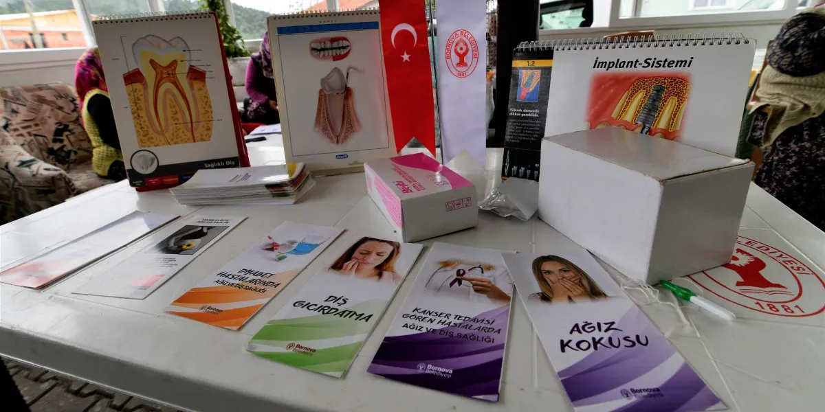 Beyaz bir masa üzerinde diş sağlığı eğitimi için hazırlanmış broşürler, bilgilendirme kartları, bir kutu ve Bornova Belediyesi logolu materyaller yer alıyor. Masanın arka kısmında diş anatomisini, implant sistemini gösteren eğitim görselleri ve bir Türk bayrağı bulunuyor. Ön tarafta ise diyabet hastalarında ağız ve diş sağlığı, diş gıcırdatma, kanser tedavisi gören hastalarda ağız ve diş sağlığı ile ağız kokusu gibi konuları içeren bilgilendirici broşürler sergileniyor.