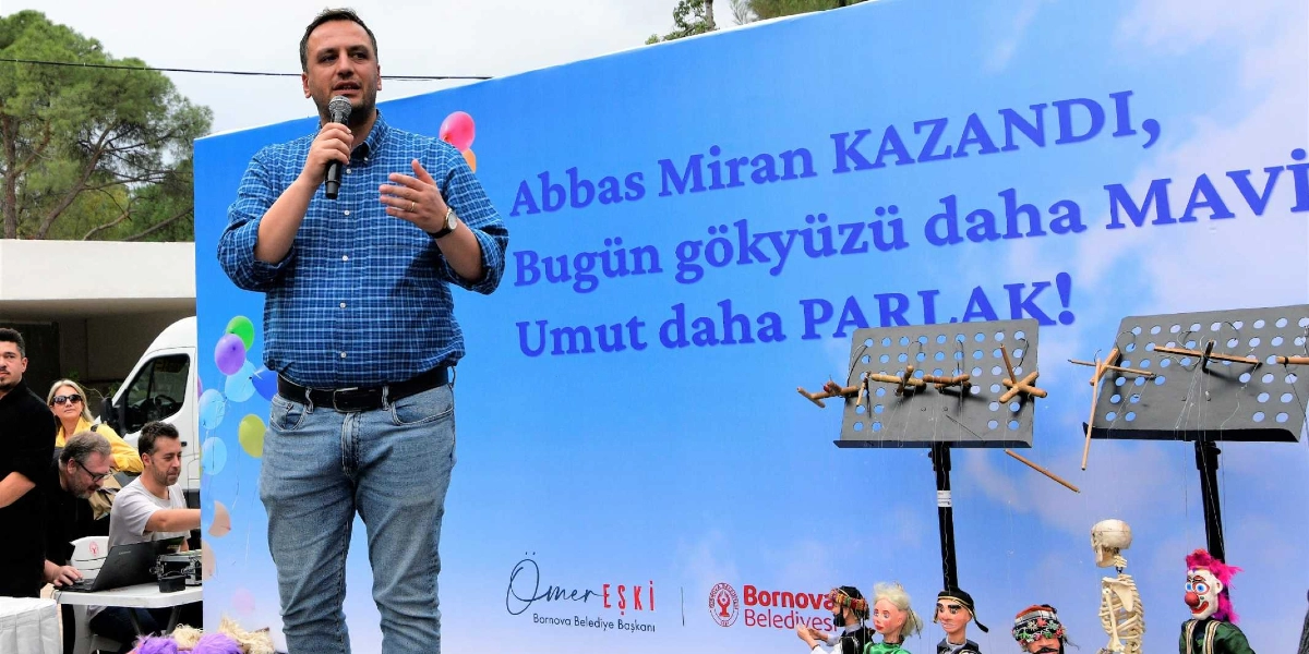 Mavi ekoseli gömlek ve kot pantolon giymiş bir erkek, elinde mikrofonla konuşma yapıyor. Arkasında üzerinde 