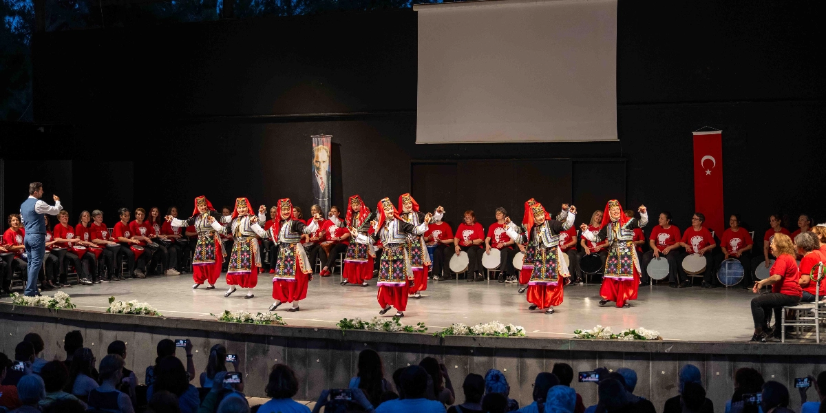 Sahne üzerinde geleneksel halk dansları kıyafetleri giymiş altı kadın dansçı, ellerinde zillerle performans sergiliyor. Dansçıların arkasında, kırmızı tişörtlü ve ellerinde darbuka tutan geniş bir müzisyen grubu oturuyor. Sahnenin solunda orkestra şefi performansın yönetimini yaparken, arka planda Atatürk posteri ve Türk bayrağı asılı. Sahne önü beyaz çiçeklerle süslenmiş olup, izleyiciler sahneyi karşıdan izliyor.