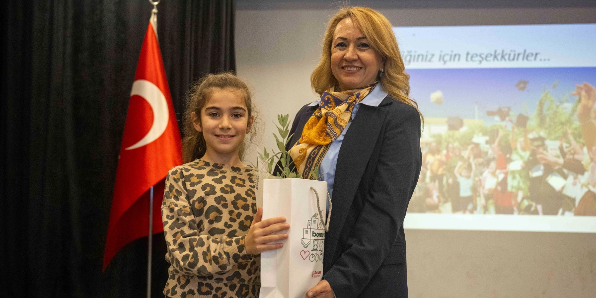 Leopar desenli kıyafet giyen genç bir kız çocuğu ile lacivert ceketli ve sarı desenli fularlı bir kadın, üzerinde Bornova Belediyesi logosu bulunan bir hediye çantasını birlikte tutarak gülümsüyor. Arka planda Türk bayrağı ve üzerinde 