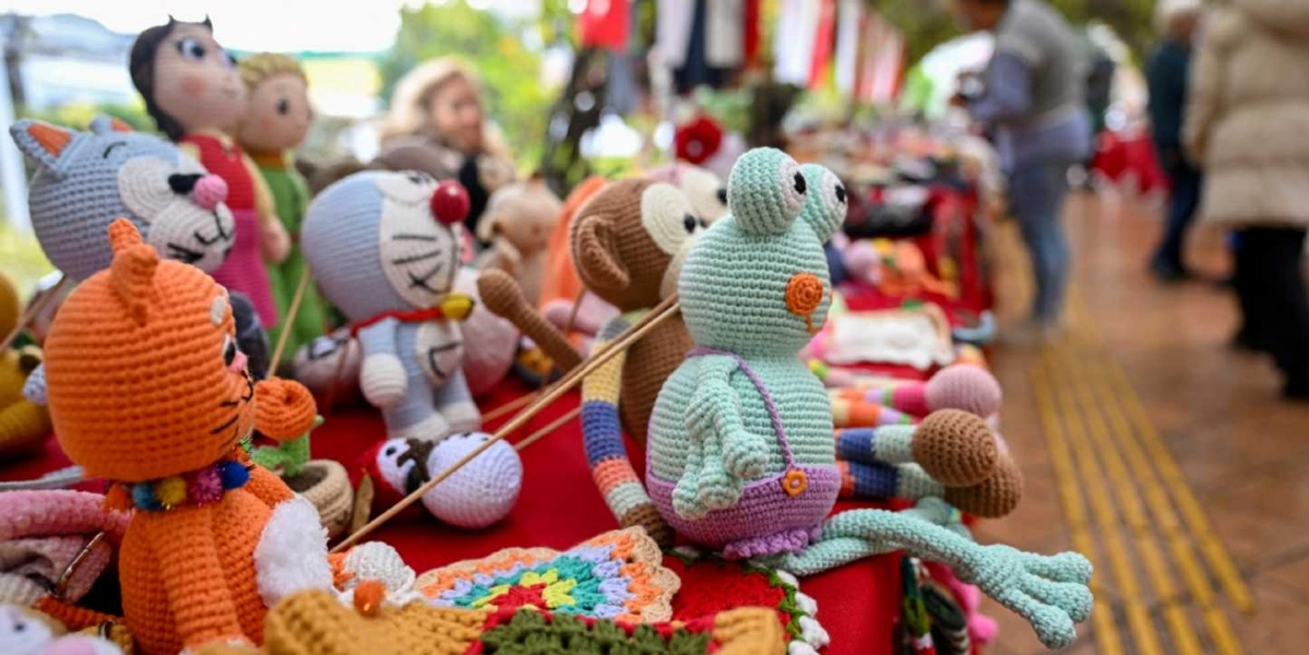 Açık havada kurulan bir stantta, kırmızı örtü üzerine dizilmiş el yapımı rengarenk amigurumi örgü oyuncaklar. Ön planda turuncu bir kedi, açık yeşil bir kurbağa, kahverengi bir maymun ve Doraemon karakteri gibi çeşitli figürler yer alıyor. Arka planda ise alışveriş yapan insanlar ve stantların devamı bulanık bir şekilde görülüyor.
