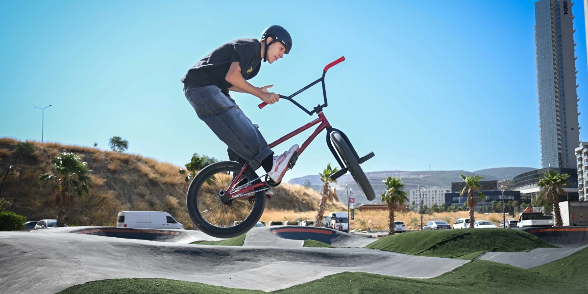 Kask takan genç bir bisikletçi, açık havada bulunan asfalt bir parkurda BMX bisikletiyle havada zıplama hareketi yapıyor. Arka planda palmiye ağaçları, tepelik bir alan ve yüksek bir bina yer alıyor.