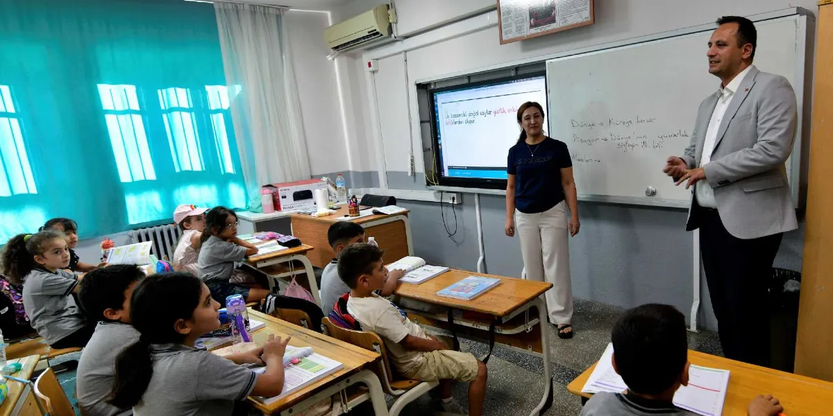 Sınıf ortamında, akıllı tahtanın önünde duran bir kadın öğretmen ve yanında konuşma yapan gri ceketli bir yetkili, sıralarda oturan ilkokul öğrencilerine hitap ediyor. Öğrenciler masalarında ders kitaplarıyla otururken, tahtada 