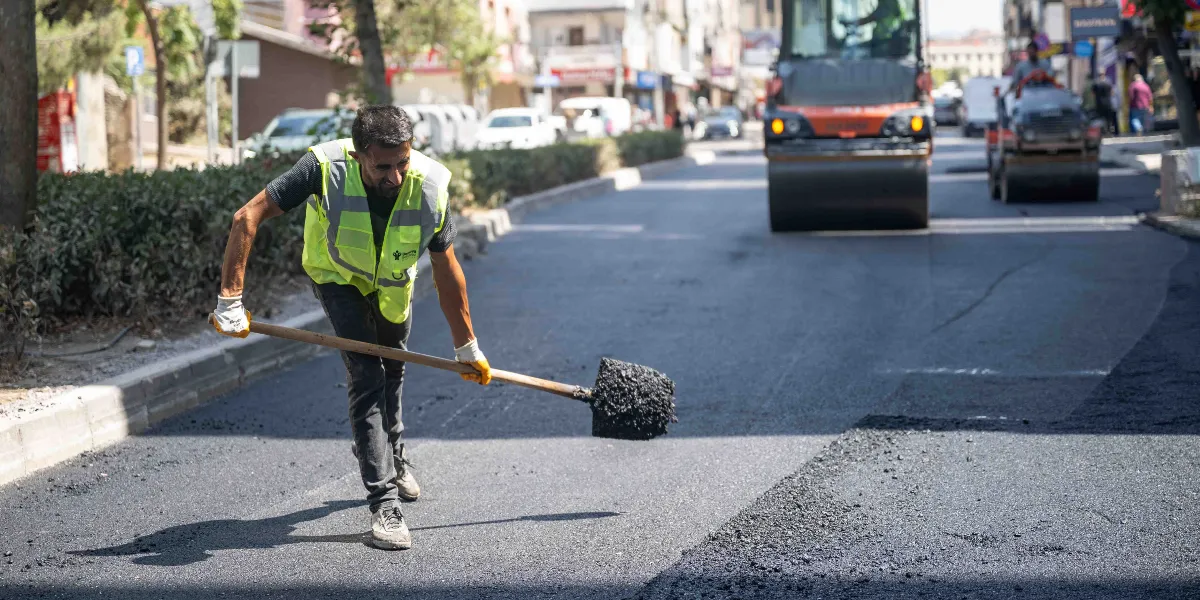 Sarı reflektörlü yelek giyen bir işçi, elindeki kürekle yeni dökülmüş asfaltı düzeltiyor. Arka planda yol çalışmasında kullanılan bir silindir ve şehir içi bir cadde manzarası yer alıyor.