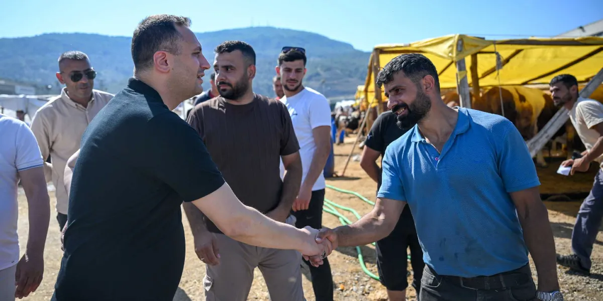 Açık havada, kurbanlık hayvan satış alanında siyah tişörtlü bir yetkili, mavi polo yaka tişört giyen bir vatandaşla tokalaşıyor. Çevredeki diğer erkekler bu anı izlerken, arka planda sarı tenteli çadırlar ve dağlık bir arazi görülüyor.