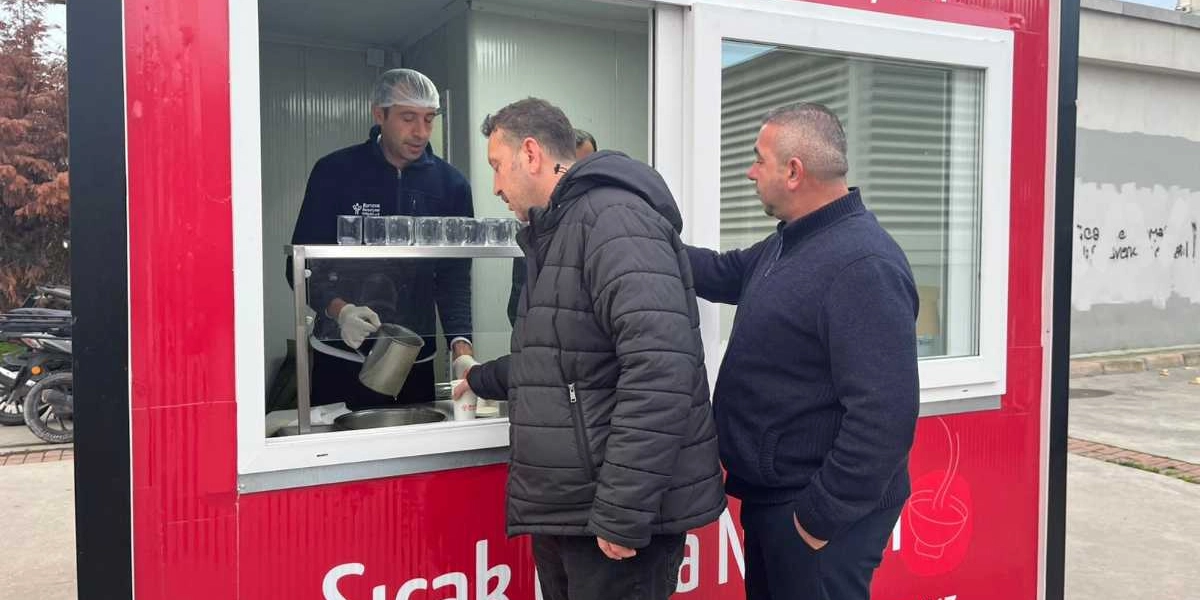Bornova Belediyesi'ne ait kırmızı renkli bir çorba dağıtım standında, bone ve eldiven takan bir görevli, standın önünde bekleyen iki kişiye içecek ikram ediyor. Standın üzerinde 