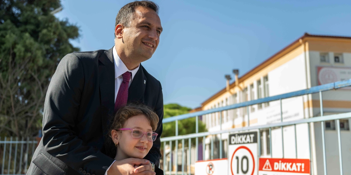 Koyu renk takım elbiseli ve bordo kravatlı bir erkek, önünde duran gözlüklü küçük bir kız çocuğunun omuzlarından tutarak gülümseyerek ileriye bakıyor. Arka planda bir okul binası ve üzerinde 