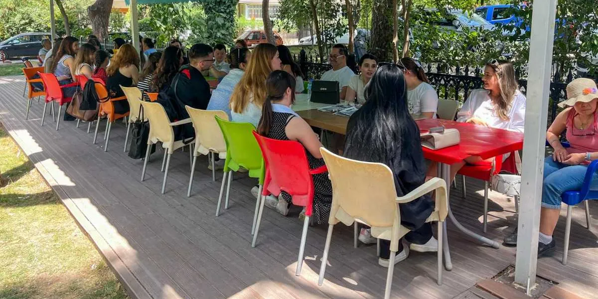 Açık havada, gölgelikli bir alanda yer alan uzun masaların etrafında toplanmış, sohbet eden bir grup genç kadın ve erkek. Katılımcılar kırmızı, sarı ve beyaz plastik sandalyelerde oturuyor, masaların üzerinde kitaplar ve kişisel eşyalar bulunuyor. Arka planda ağaçlar ve park edilmiş araçlar görülüyor.
