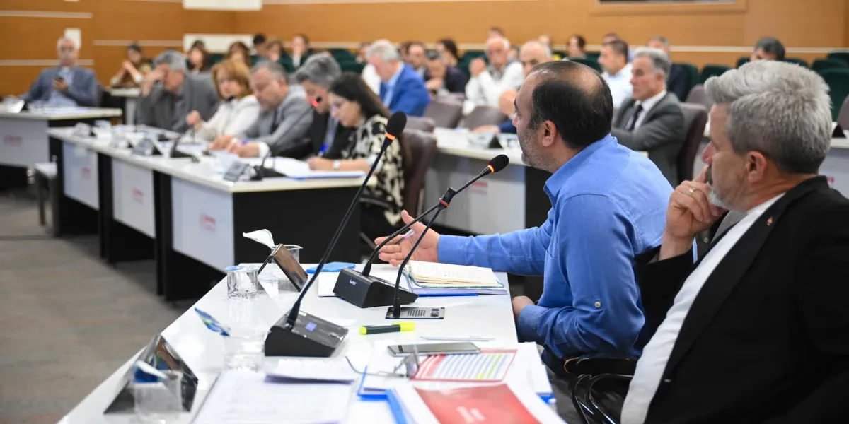 Bornova Belediye Meclis toplantısında, mavi gömlekli bir konuşmacı önündeki mikrofonla hitap ederken, yanında siyah ceketli bir katılımcı onu dinliyor. Toplantı salonunda sıralar halinde oturan çok sayıda meclis üyesi, önlerindeki evraklar ve dosyalarla oturumu takip ediyor.
