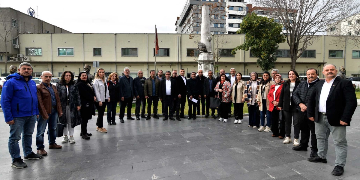 Bornova Muhtarlar Akademisi kapsamında, dış mekanda, arkalarında uzun bir anıt ve bina bulunan geniş bir grup muhtar ve katılımcı yan yana dizilmiş şekilde duruyor. Çoğunluğu resmi ve yarı resmi kıyafetler giymiş olan grup, kameraya bakarak poz veriyor.