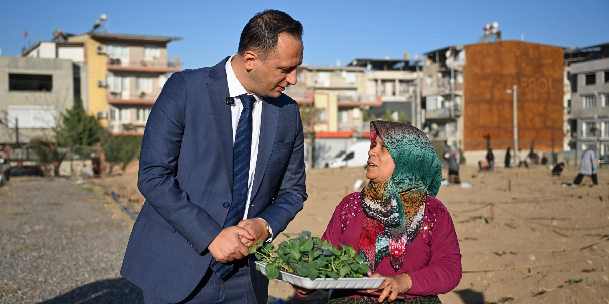 Lacivert takım elbiseli bir erkek, elinde fide tepsisi tutan başörtülü bir kadınla açık bir arazide sohbet ediyor. Arka planda yerleşim yerleri ve toprakla uğraşan diğer insanlar görülüyor.