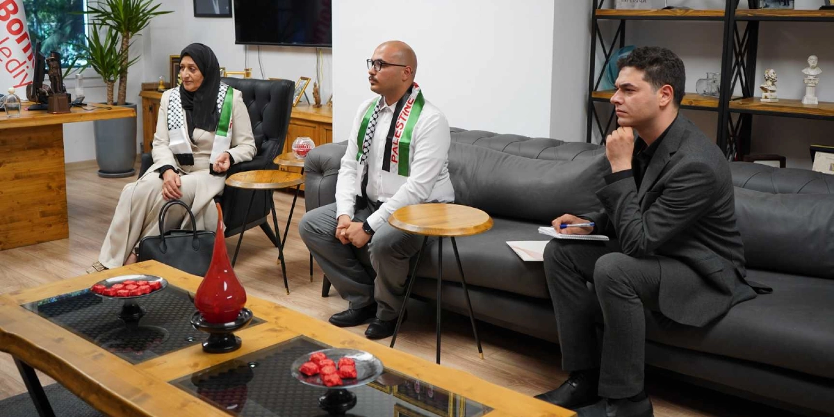 Ofis ortamında bir araya gelen üç kişi, gri bir koltukta ve tekli koltukta oturuyor. Solda, siyah başörtülü ve üzerinde Filistin bayrağı desenli atkı bulunan bir kadın, ortada beyaz gömlekli ve yine Filistin bayrağı desenli atkı takan gözlüklü bir erkek, sağda ise koyu renk takım elbiseli, elinde not defteri ve kalem tutan bir erkek yer alıyor. Önlerinde ahşap bir orta sehpa üzerinde ikramlıklar bulunuyor.