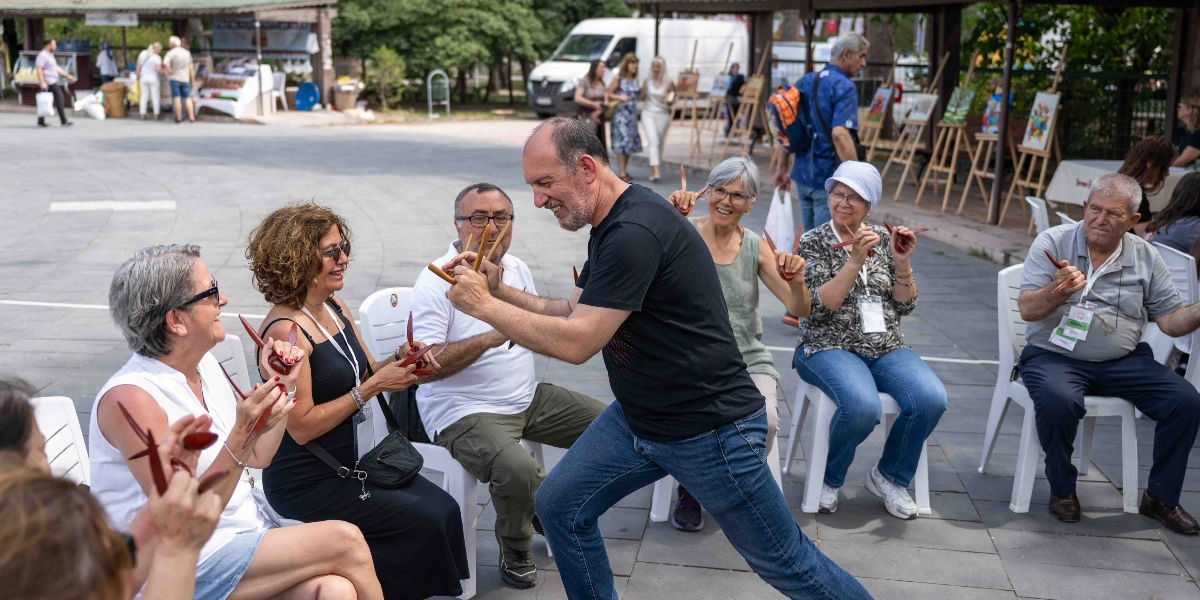 Açık havada, park benzeri bir alanda bir grup yetişkin, ellerinde tuttukları ahşap kaşıkları birbirine vurarak ritim tutuyor. Ortada siyah tişörtlü bir adam neşeyle kaşık çalarak dans ederken, çevresindeki beyaz plastik sandalyelerde oturan katılımcılar ona eşlik ediyor. Arka planda resim şövaleleri ve yürüyen insanlar görülüyor.