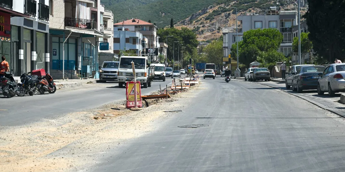 Pınar Caddesi'nde devam eden yol yenileme çalışmaları nedeniyle caddenin ortasında kazılmış bir kanal ve üzerinde uyarı levhası bulunuyor. Trafiğin aktığı caddenin kenarlarında park etmiş araçlar, motosikletler ve dükkanlar yer alırken, arka planda ağaçlıklı bir yamaç ve binalar görülüyor.