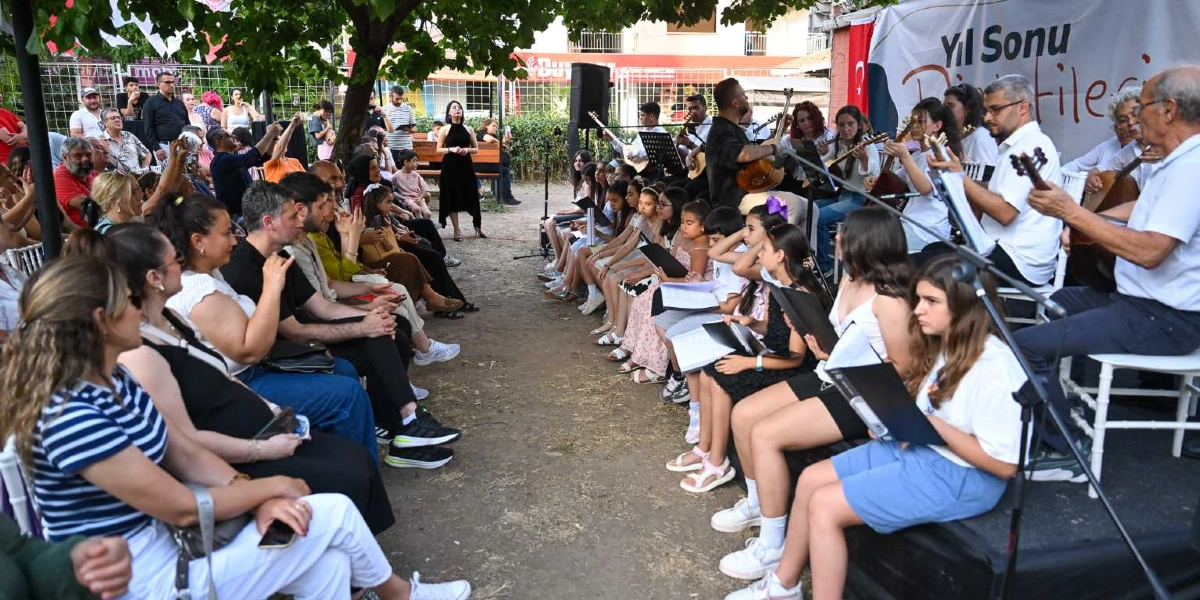 Açık havada, ağaçların altında düzenlenen yıl sonu dinletisinde, bir grup genç müzisyen ellerinde notalarla sandalyelerde oturarak performans sergiliyor. Yanlarında gitar çalan yetişkinler ve eğitmenler eşlik ederken, izleyiciler de karşı tarafta sandalyelerde oturarak konseri ilgiyle takip ediyor. Arka planda üzerinde 