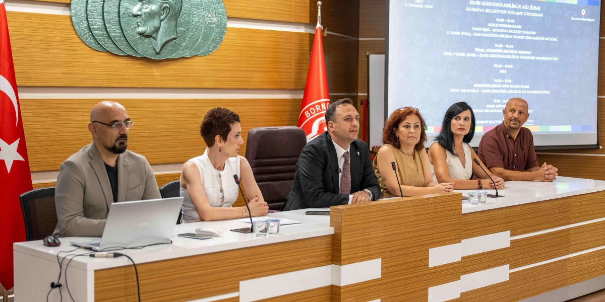 Bornova Belediyesi'nde düzenlenen İzmir Sürdürülebilirlik Ağı toplantısında, uzun bir masa etrafında yan yana oturan altı kişilik bir panelist grubu yer alıyor. Masanın arkasındaki duvarda Türk bayrağı, Bornova Belediyesi bayrağı ve Atatürk rölyefi bulunuyor. Panelistlerin arkasındaki projeksiyon ekranında ise toplantı programı ve konuşmacı isimleri yer alıyor.