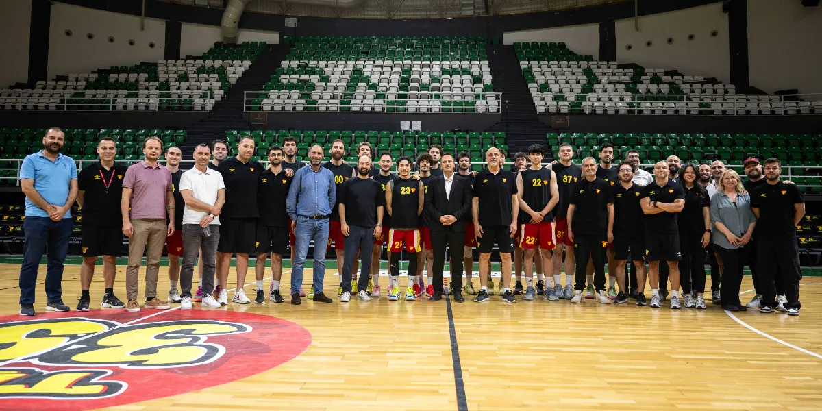 Bornova Belediye Başkanı Ömer Eşki, Göztepe Hentbol takımı oyuncuları ve teknik ekibiyle birlikte kapalı spor salonunda yan yana durarak toplu fotoğraf çektiriyor. Oyuncuların bir kısmı Göztepe logolu siyah antrenman formaları ve şortlarıyla yer alırken, teknik ekip ve Başkan Eşki günlük kıyafetleriyle bulunuyor. Arka planda yeşil ve beyaz koltuklu boş tribünler görülüyor.