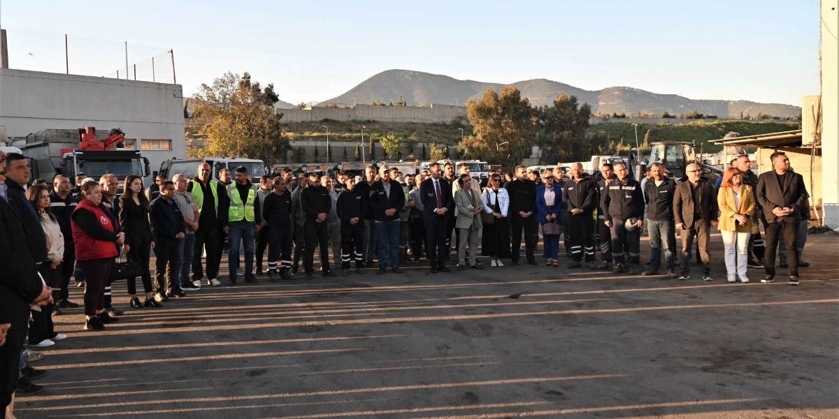 Açık havada, asfalt bir alanda yan yana dizilmiş, çoğunluğu erkeklerden oluşan kalabalık bir grup insan duruyor. Arka planda iş makineleri, kamyonlar ve uzakta dağ manzarası görülüyor. Gün batımına yakın bir saatte çekilen fotoğrafta, insanların uzun gölgeleri yere düşüyor. Gruptakilerin bir kısmı günlük kıyafetlerle, bir kısmı ise iş kıyafetleri veya resmi giysilerle yer alıyor.